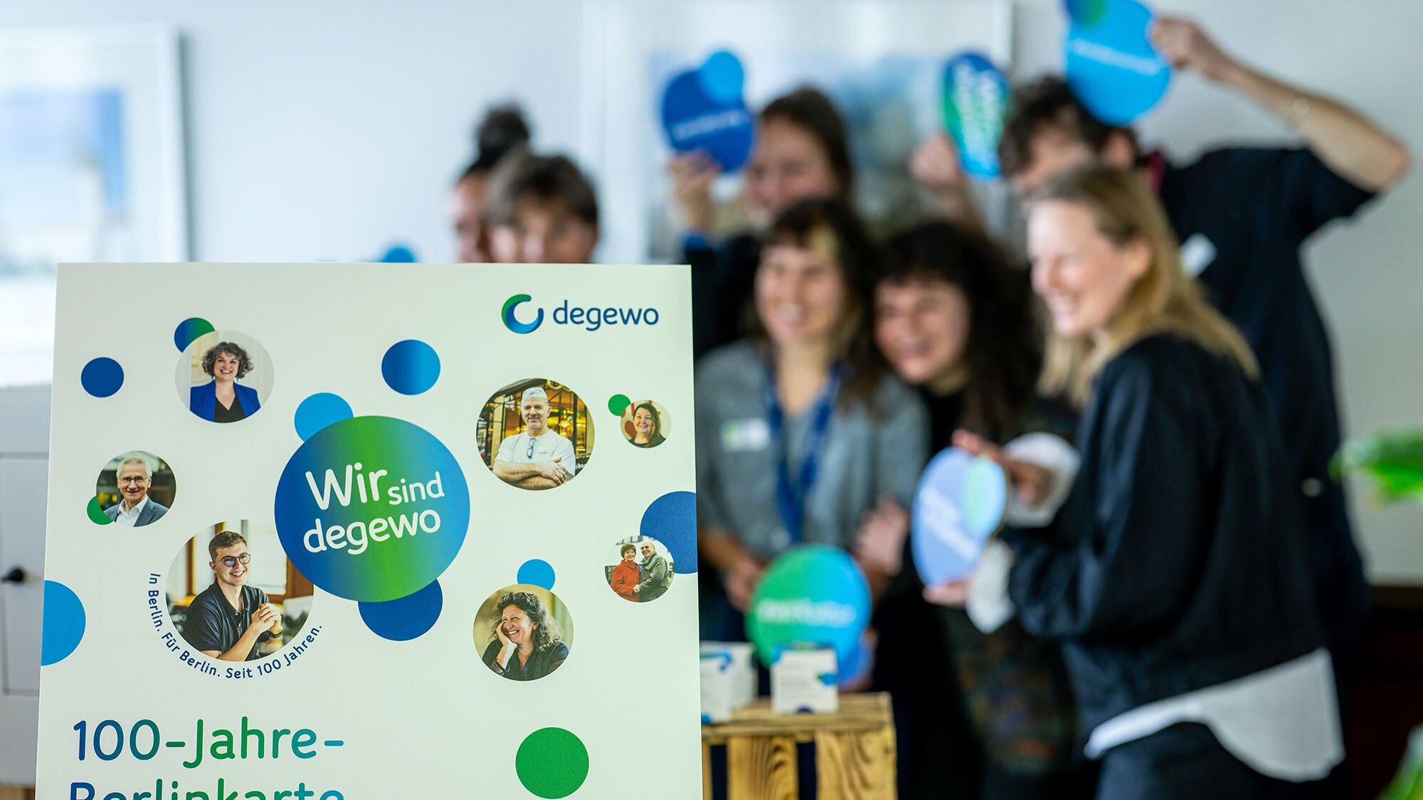 Im Vordergrund steht ein Aufsteller mit der Aufschrift „Wir sind degewo“ und „100-Jahre-Berlinkarte“ sowie mehreren Porträtfotos. Im Hintergrund posiert eine Gruppe von Personen mit runden Schildern in Blau- und Grüntönen. Die Personen sind unscharf dargestellt, während der Aufsteller im Fokus steht.