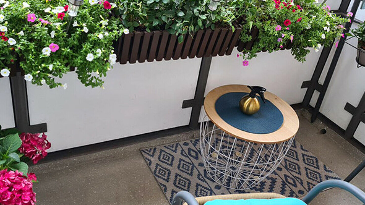 ein mit vielen kleinen Topfpflanzen dekorierter Balkon. Auf dem Balkon steht ein kleiner Tisch und ein Stuhl mit blauem Kissen.