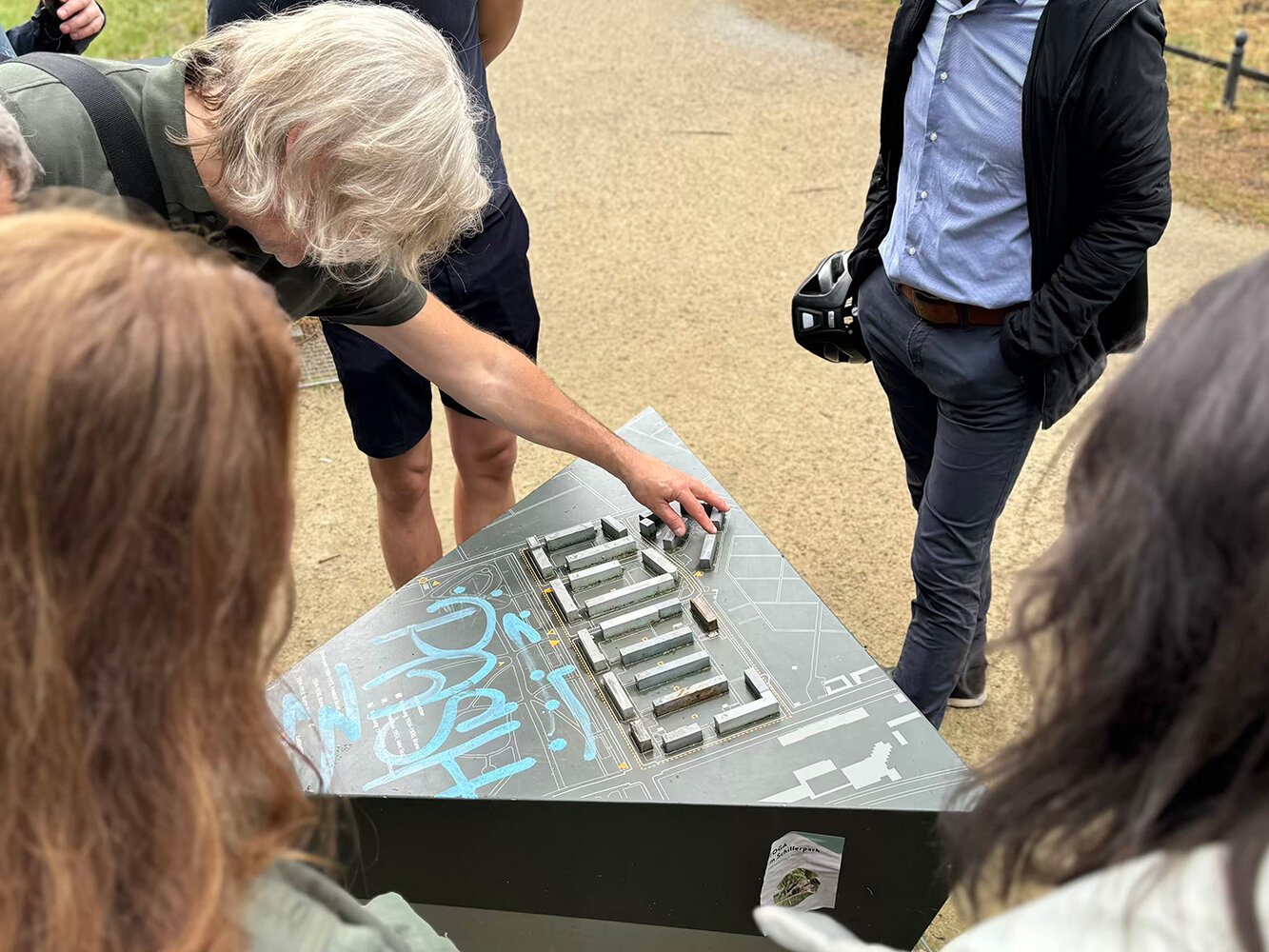 Mehrere Personen stehen im Freien um ein Modell einer Wohnanlage während ein Mann auf einzelne Gebäude zeigt und die Planung erklärt.