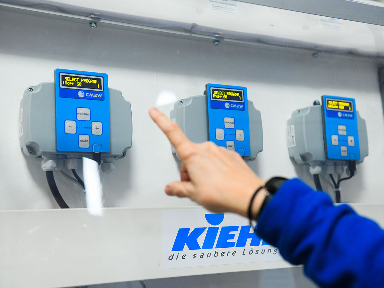 Eine Hand zeigt auf ein digitales Bedienfeld eines blauen Dosiergeräts für Reinigungsprogramme das an der Wand montiert ist.