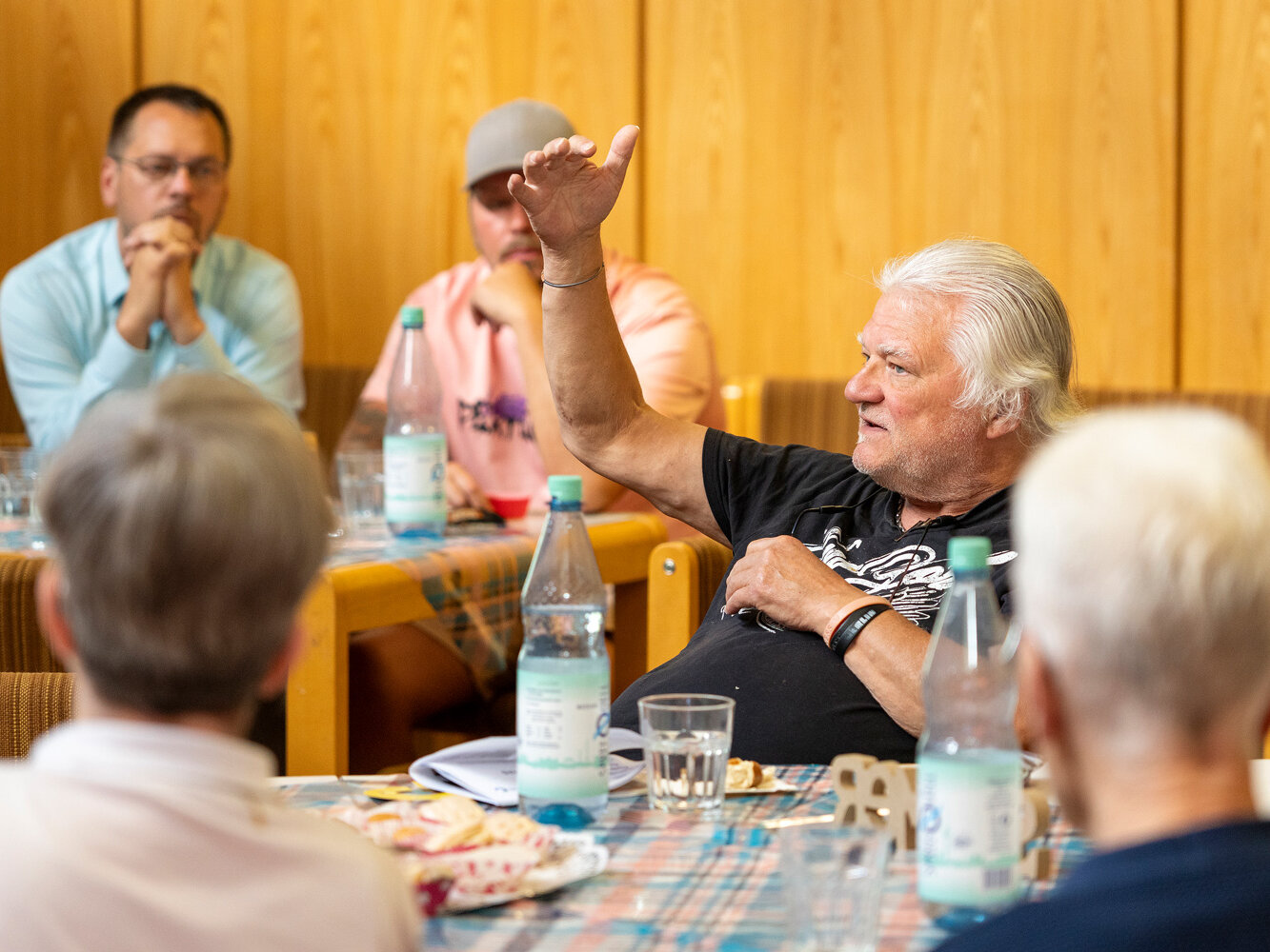 Ein älterer Bewohner meldet sich während einer Diskussionsrunde zu Wort. Mehrere Teilnehmende sitzen an Tischen und hören aufmerksam zu.