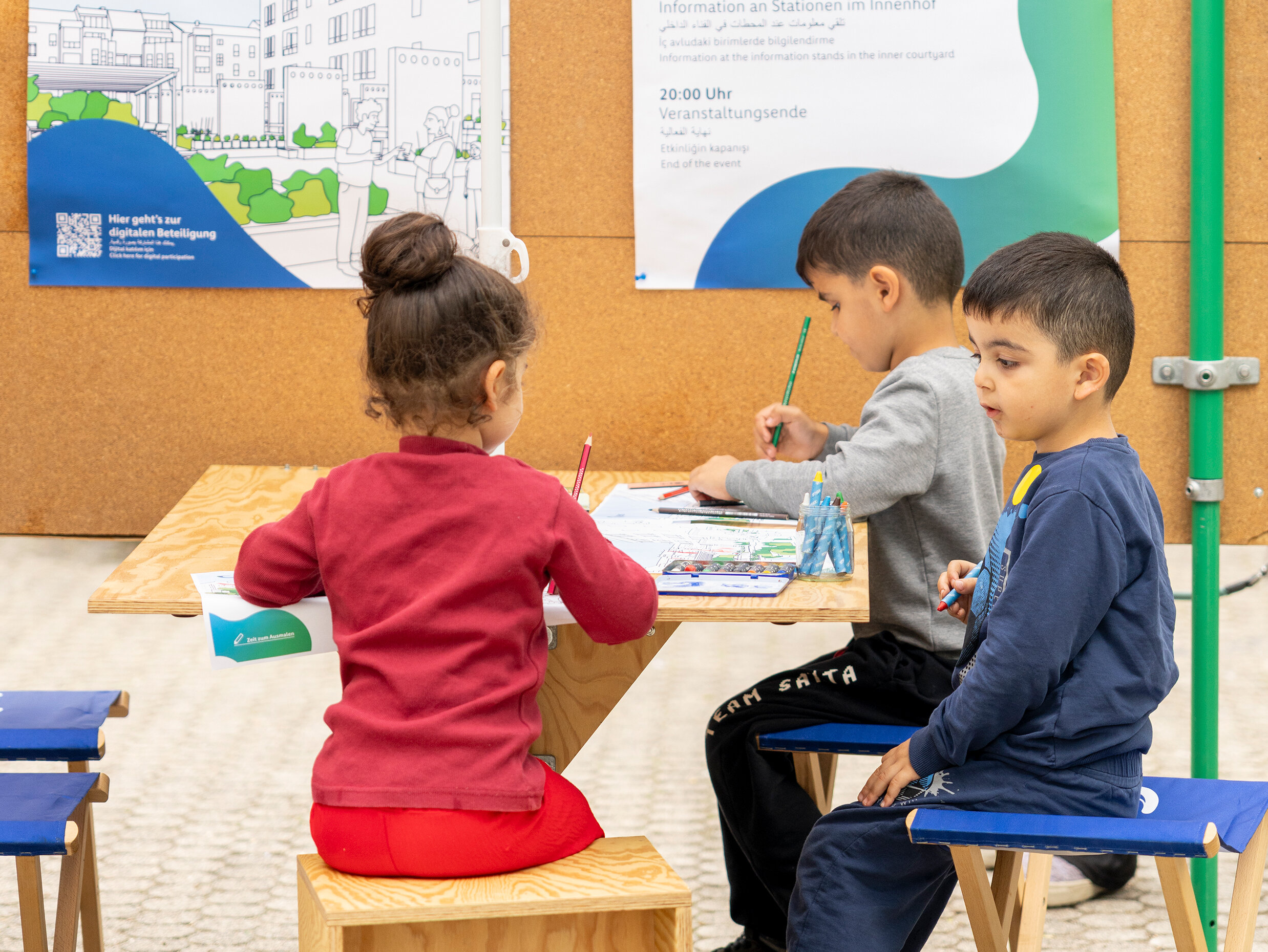 Drei Kinder sitzen an einem Tisch und malen. Hinter dem Tisch eine große Korkwand mit zwei degewo-Plakaten. Über den Kindern ein Sonnenschirm mit degewo-Logo.