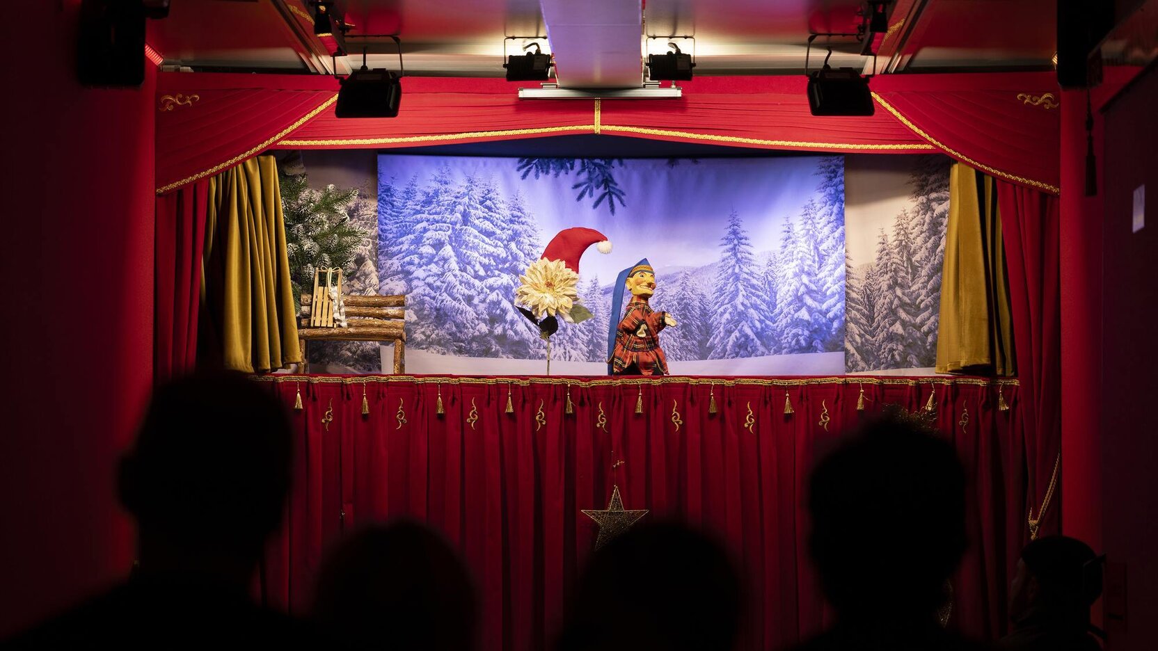 Puppentheater auf einer weihnachtlich dekorierten Bühne mit winterlicher Waldkulisse und Publikum im Vordergrund.