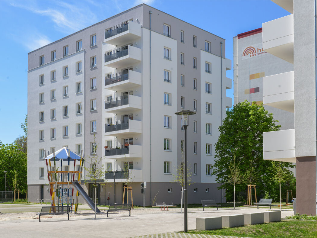 Außenansicht degewo-Neubau Eugen-Roth-Weg 4 bis 10 mit Spielplatz und Straßenlaterne neben Sitzgelegenheiten im Vordergrund.