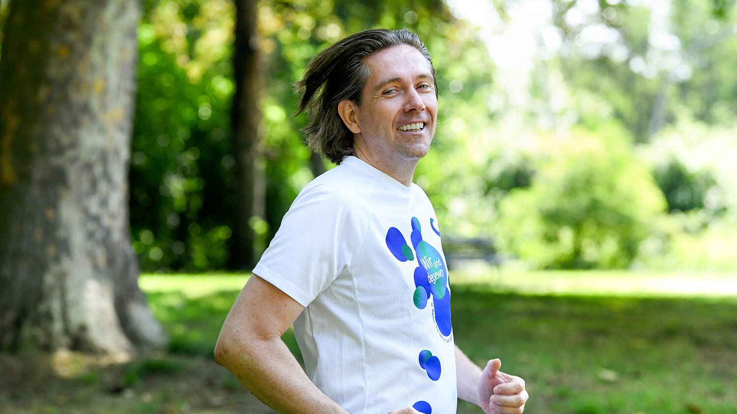Ein Mann joggt in einem Park in Berlin. Dabei lächelt er in die Kamera.
