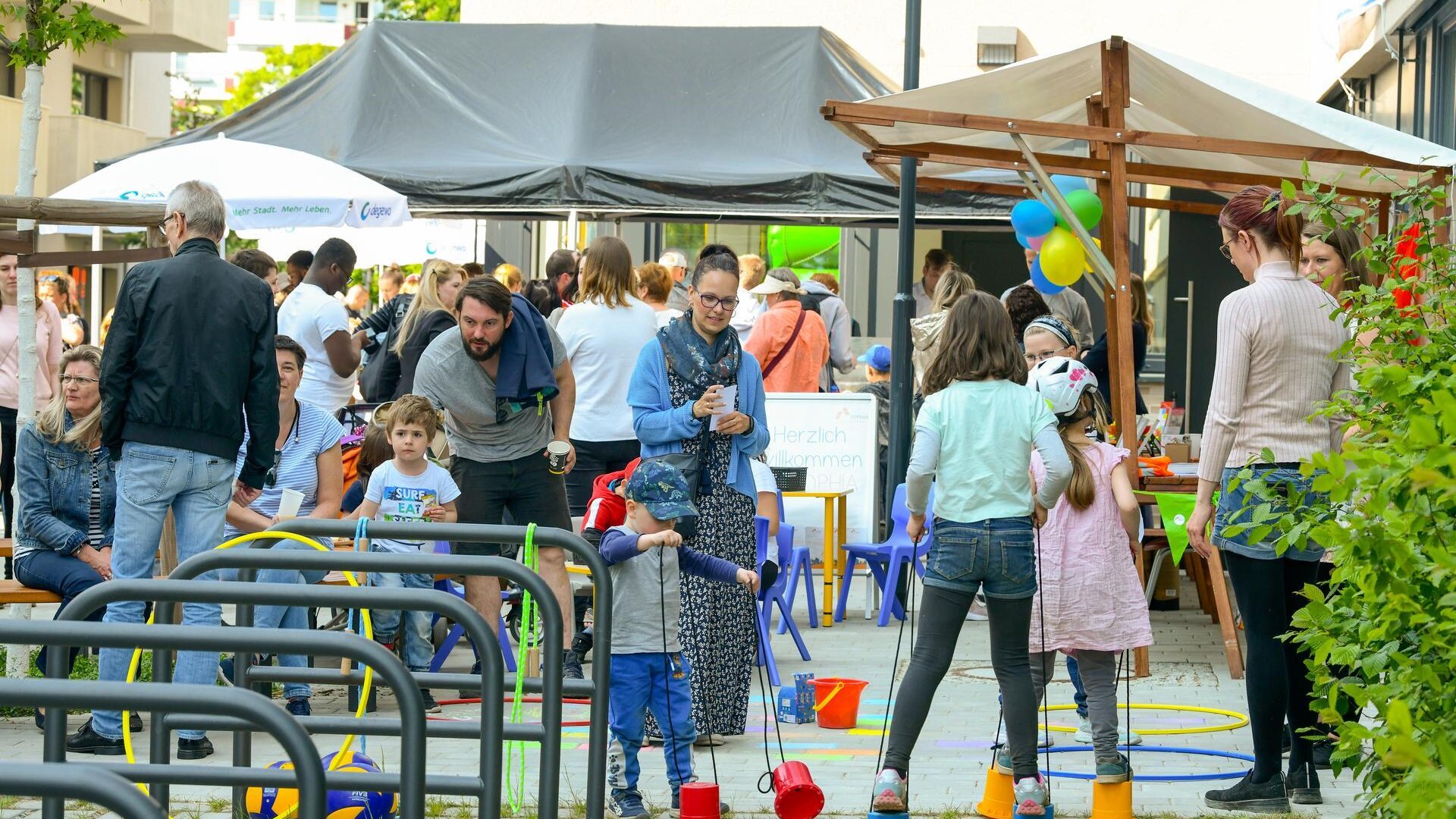 Kinder und Erwachsene besuchen ein Nachbarschaftsfest mit Spielangeboten im Innenhof einer Wohnanlage.