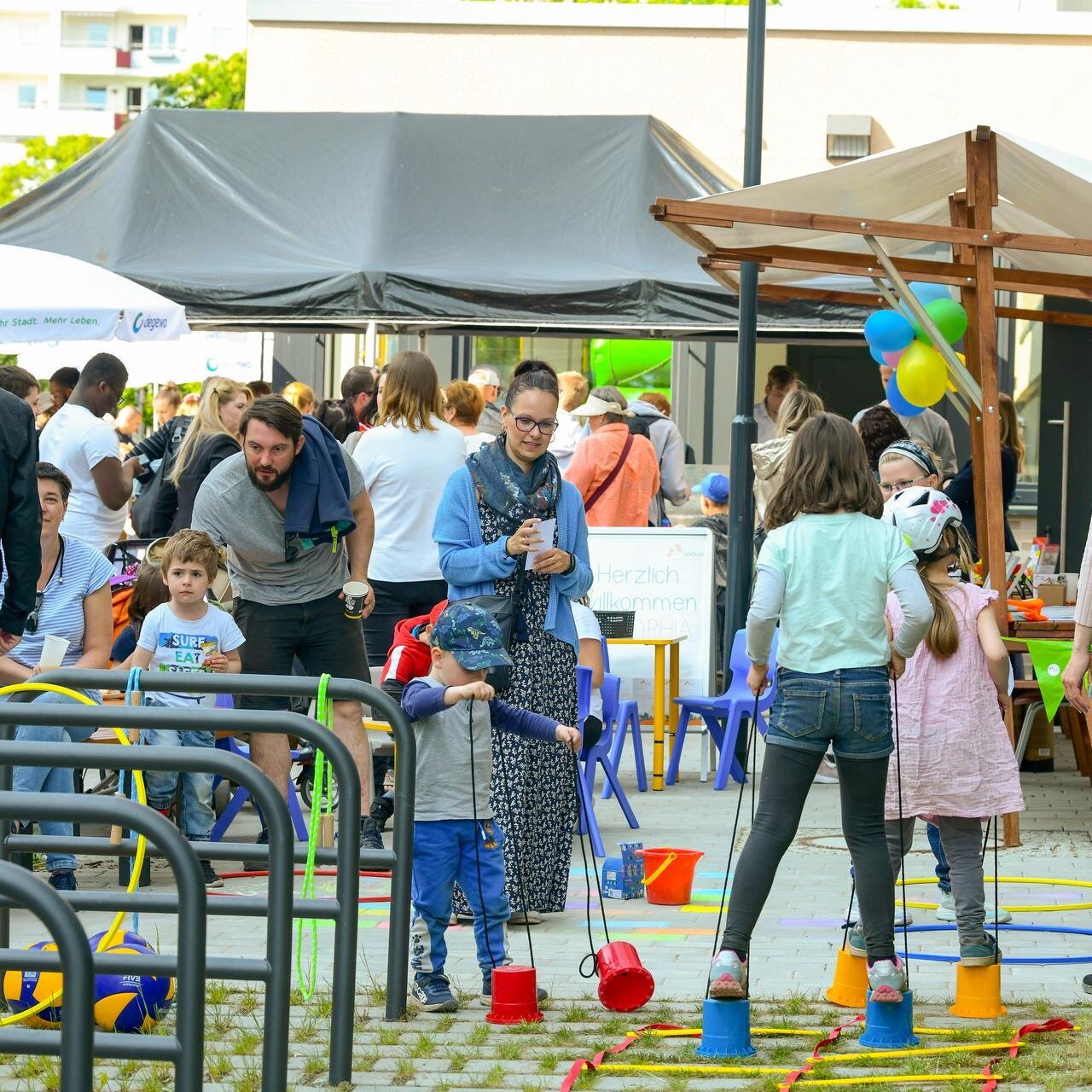 Kinder und Erwachsene besuchen ein Nachbarschaftsfest mit Spielangeboten im Innenhof einer Wohnanlage.