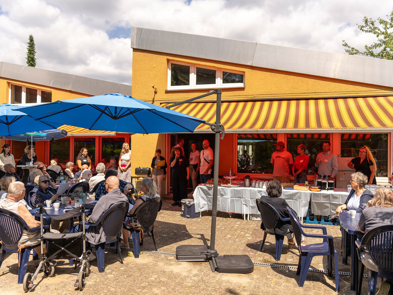 Blick auf den Außenbereich des Sanierungscafés: Viele Bewohnerinnen und Bewohner sitzen an Tischen unter Sonnenschirmen vor dem Gemeinschaftshaus.