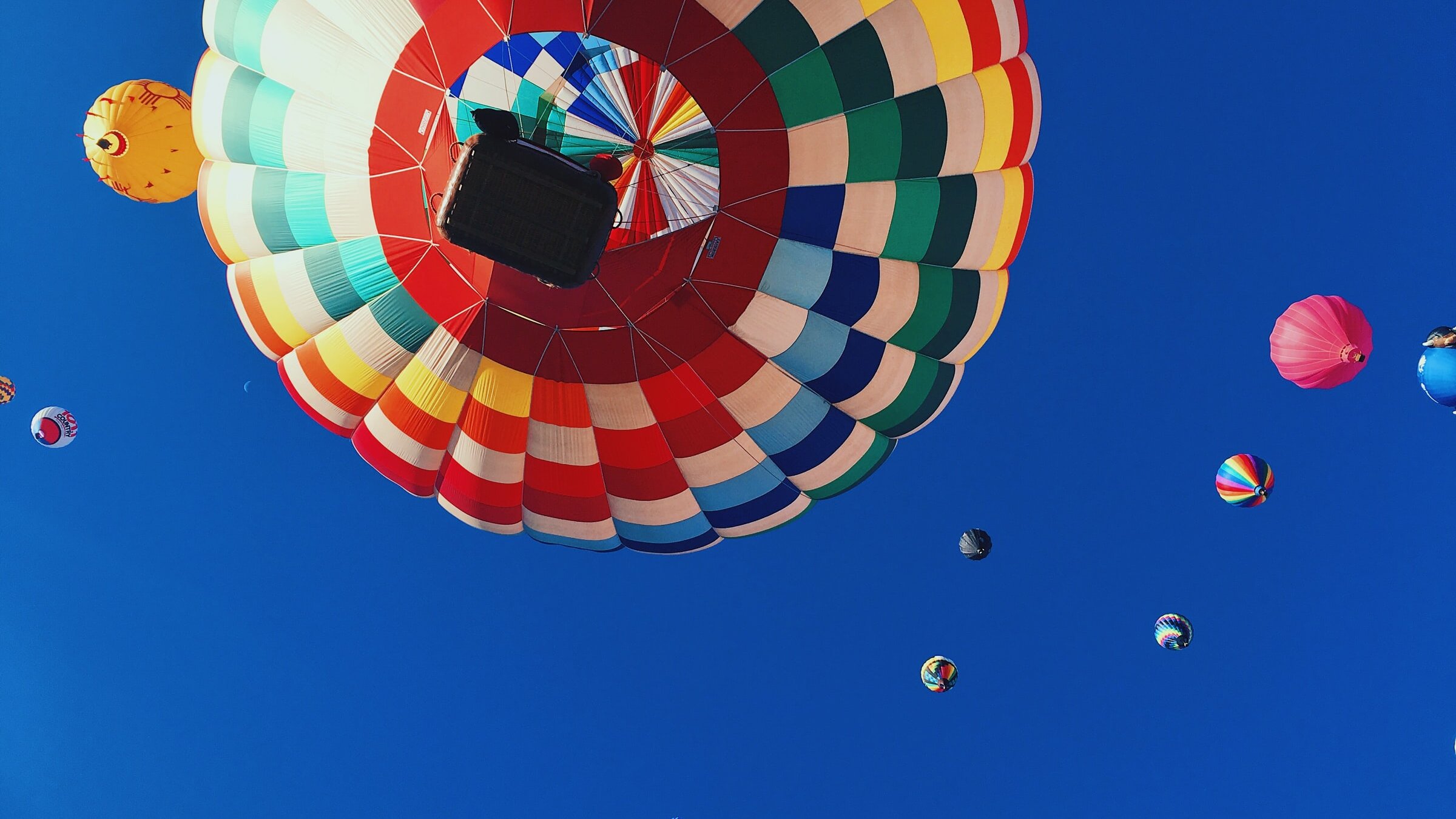 Blick von unten auf mehrere Heißluftballons am Himmel; im Vordergrund ist die Unterseite eines großen, mehrfarbig gestreiften Ballons mit Korb zu sehen, weitere kleinere Ballons schweben vor einem klaren, blauen Himmel.