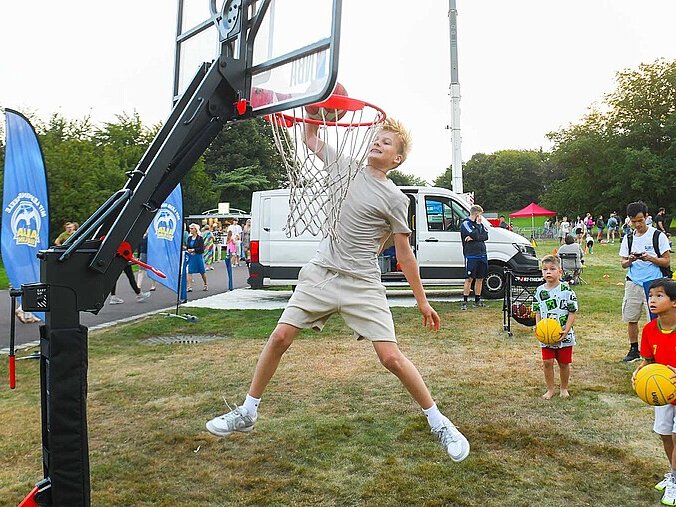 Eine Junge springt an einem Basketballkorb in die Höhe. Andere Kinder jubeln ihm dabei zu.