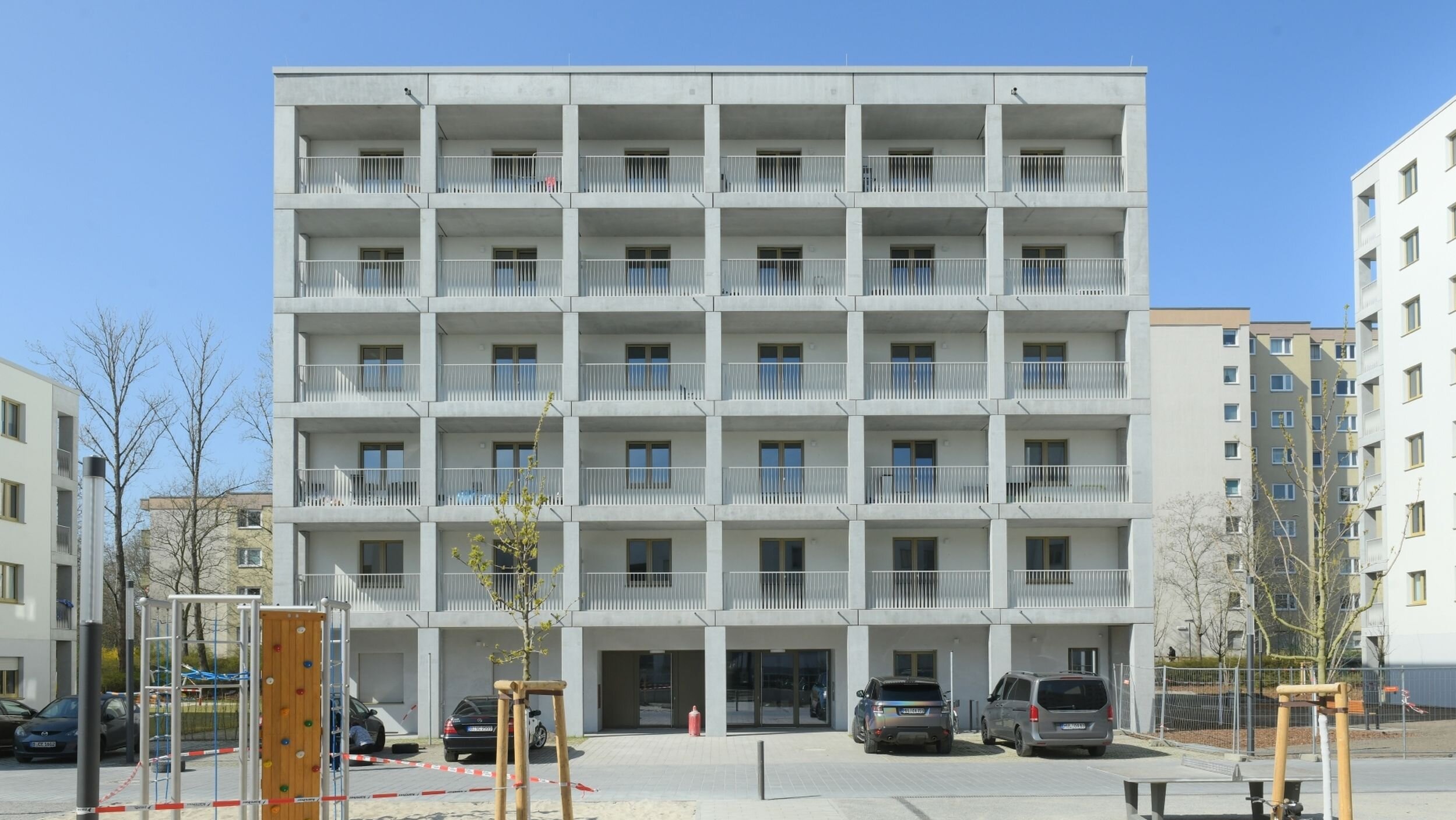 Außenansicht degewo-Neubau Theodor-Loos-Weg 31 bis 35 mit Spielplatz, Straßenlaterne, neu gepflanzten Bäumen, Weg und Tischtennisplatte im Vordergrund.