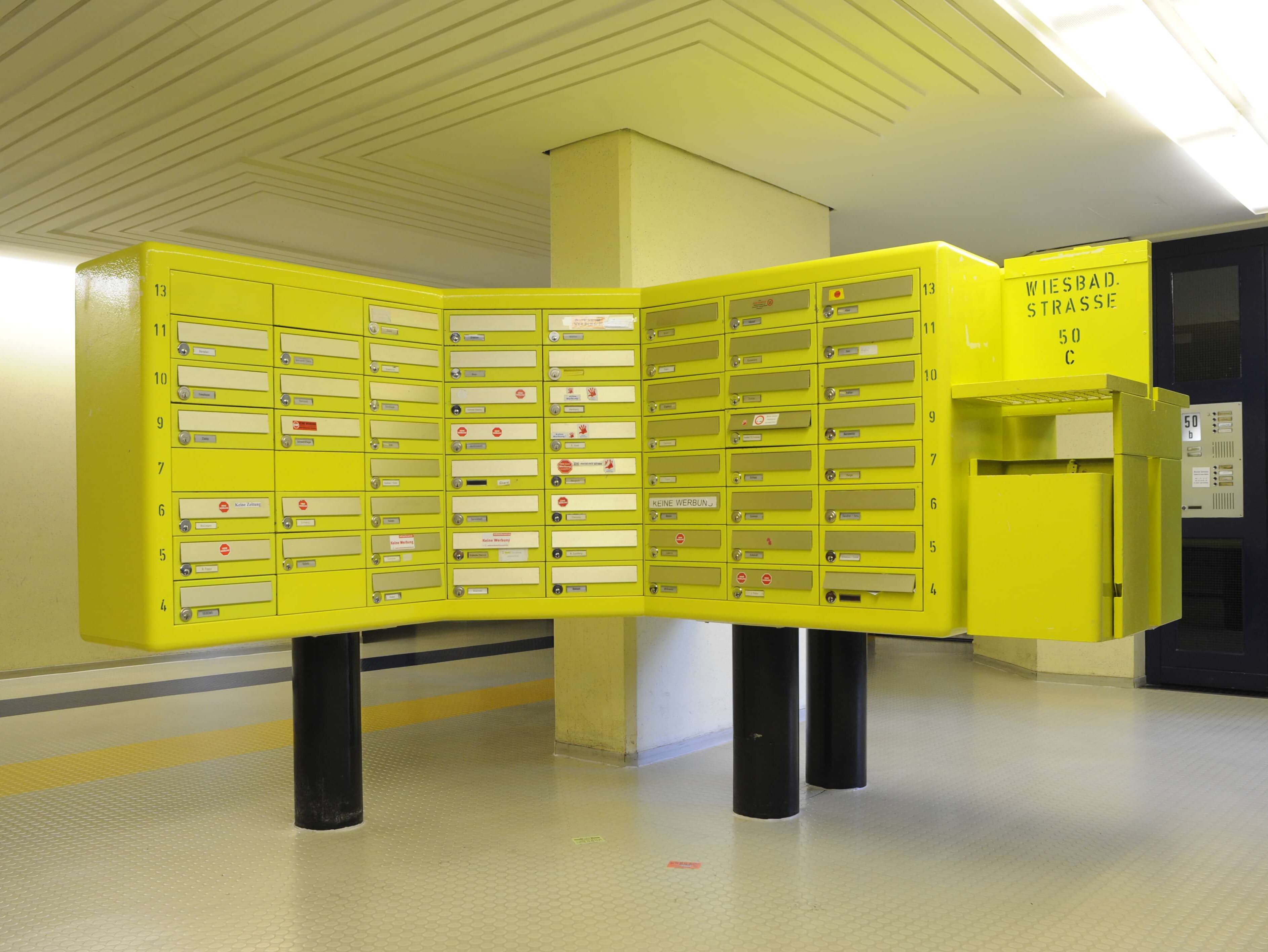 Leuchtend gelbe, großformatige Briefkastenanlage im Eingangsbereich der Wohnanlage „Die Schlange“, montiert auf schwarzen Säulen.
