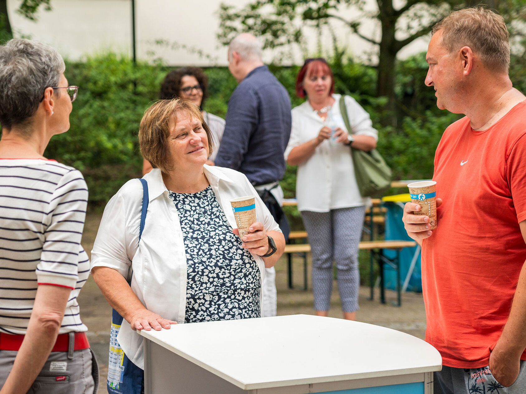 Drei Personen stehen um einen Tisch und unterhalten sich. Im Hintergrund Sitzbänke und weitere Personen. 