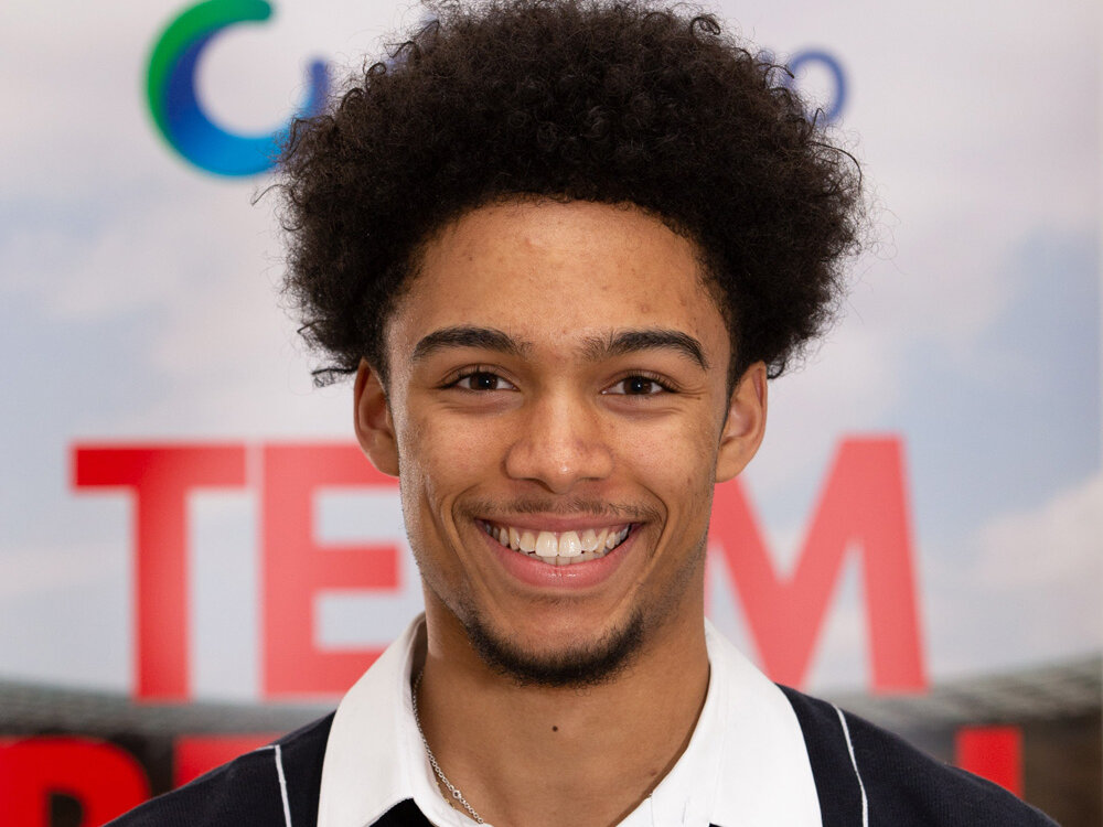 Ein junger Mann steht lächelnd vor einem Hintergrund mit den Logos von LOTTO Berlin und degewo. Er trägt ein weißes Hemd und eine dunkle Strickweste.