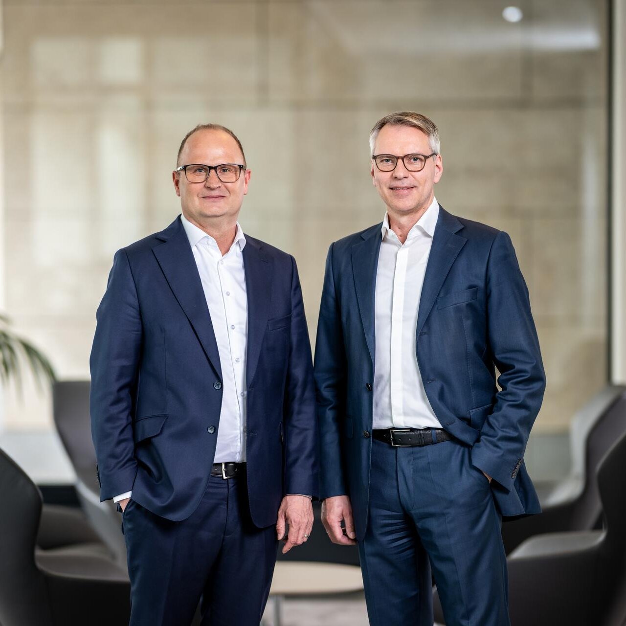 Die beiden Vorstandsmitglieder Kai-Marten Maack und Pascal Atzert stehen vor einer Glasscheibe um eine Sitzgruppe herum und lachen in die Kamera. Sie tragen Anzug. 