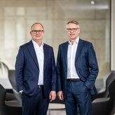 Die beiden Vorstandsmitglieder Kai-Marten Maack und Pascal Atzert stehen vor einer Glasscheibe um eine Sitzgruppe herum und lachen in die Kamera. Sie tragen Anzug. 