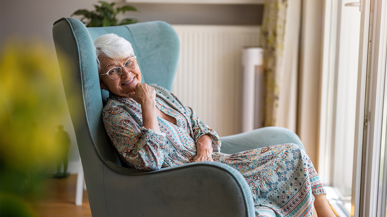 Eine ältere Frau sitzt auf einem Stuhl vor dem Fenster und lächelt.