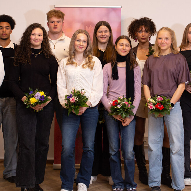 Gruppenfoto mehrerer junger Athletinnen und Athleten, die in einem Innenraum nebeneinander stehen und Blumensträuße halten. Rechts steht eine erwachsene Person in einem roten Blazer.