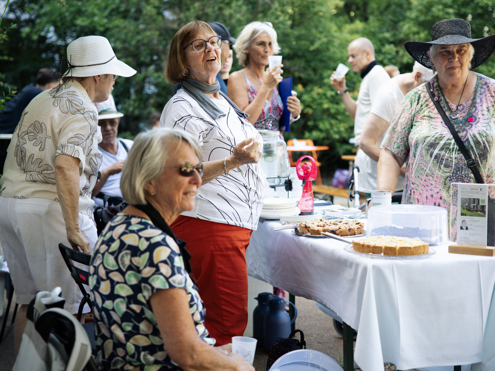 Eine Gruppe älterer Menschen steht beisammen. Manche unterhalten sich, andere schauen in die Ferne. Vor ihnen ist ein Tisch aufgestellt. Darauf steht Kuchen. 