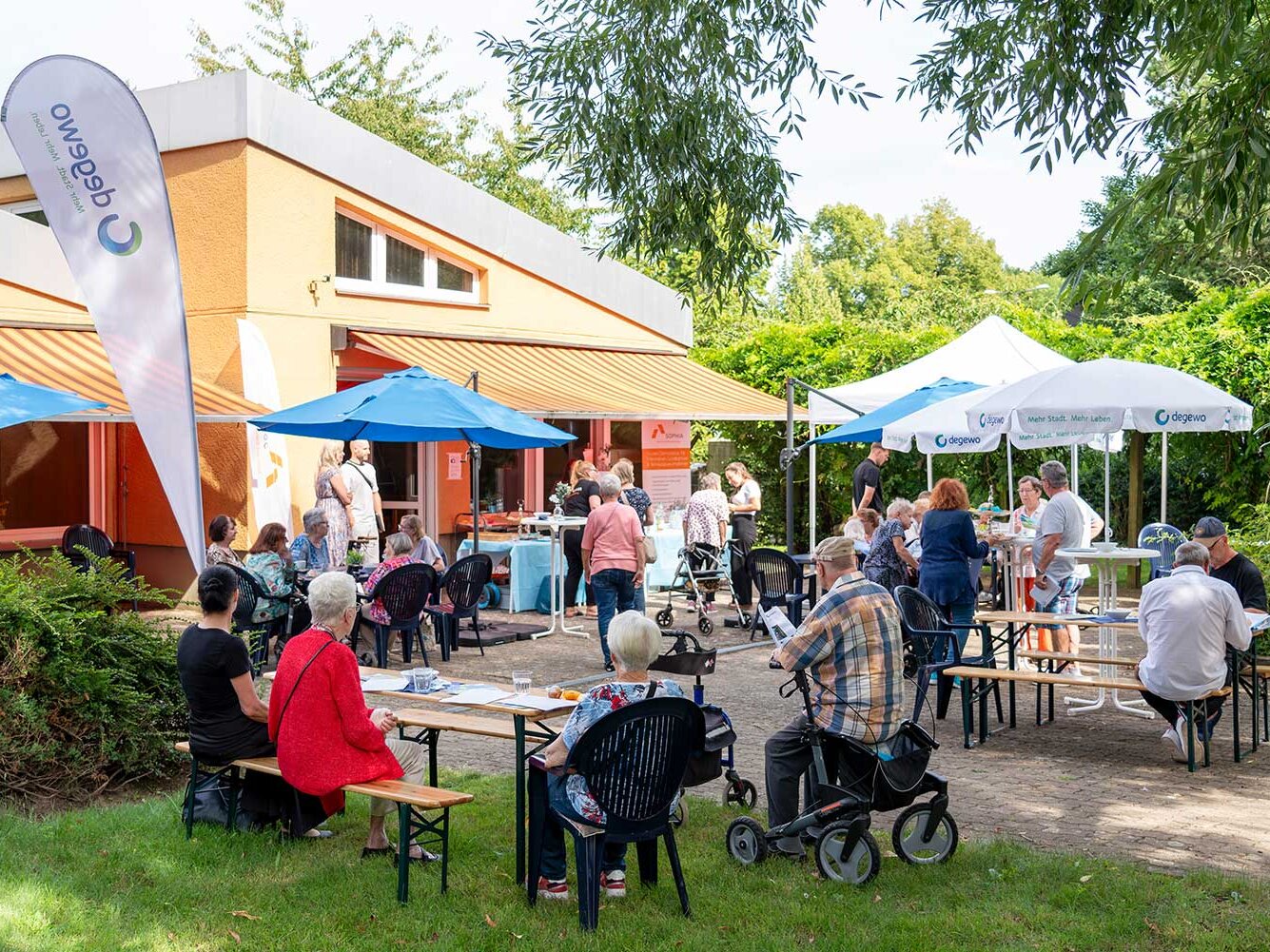 Menschen sitzen im Garten vor einem Gemeinschaftshaus bei einer Informationsveranstaltung. Unter blauen und weißen Sonnenschirmen kommen Bewohnerinnen und Bewohner miteinander ins Gespräch.