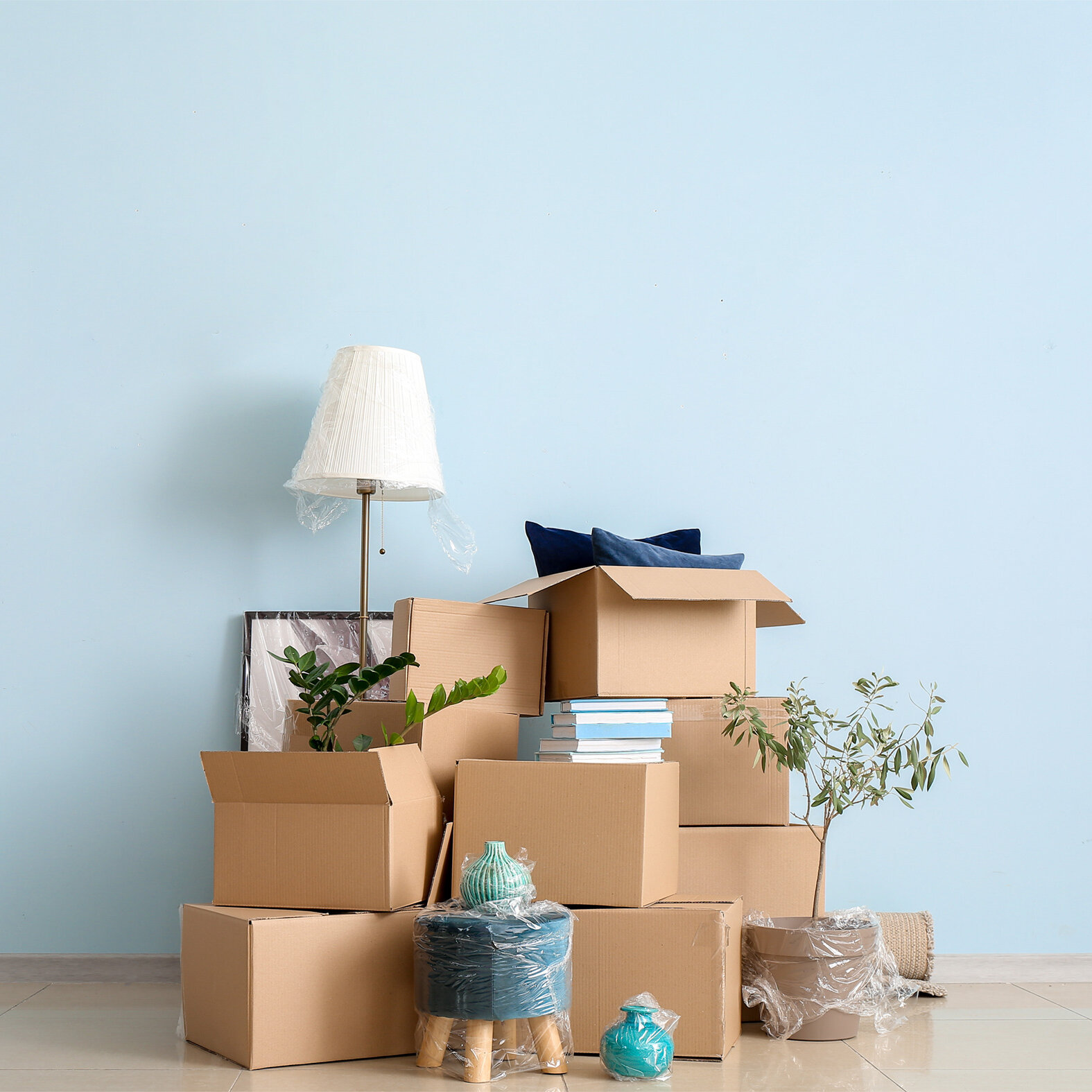 Mehrere Umzugskartons sind vor einer hellblauen Wand gestapelt. Zwischen den Kartons stehen eine verpackte Stehlampe, Zimmerpflanzen, Bücher und dekorative Gegenstände.