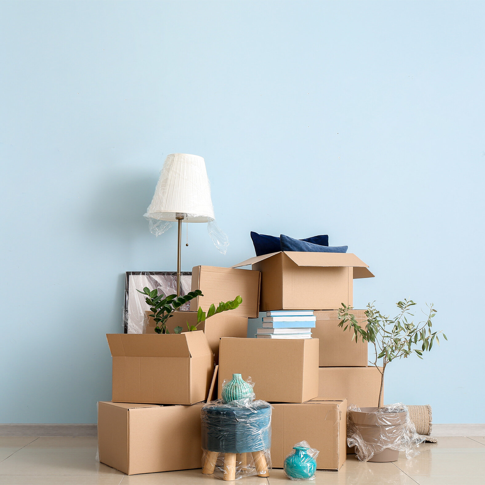 Mehrere Umzugskartons sind vor einer hellblauen Wand gestapelt. Zwischen den Kartons stehen eine verpackte Stehlampe, Zimmerpflanzen, Bücher und dekorative Gegenstände.