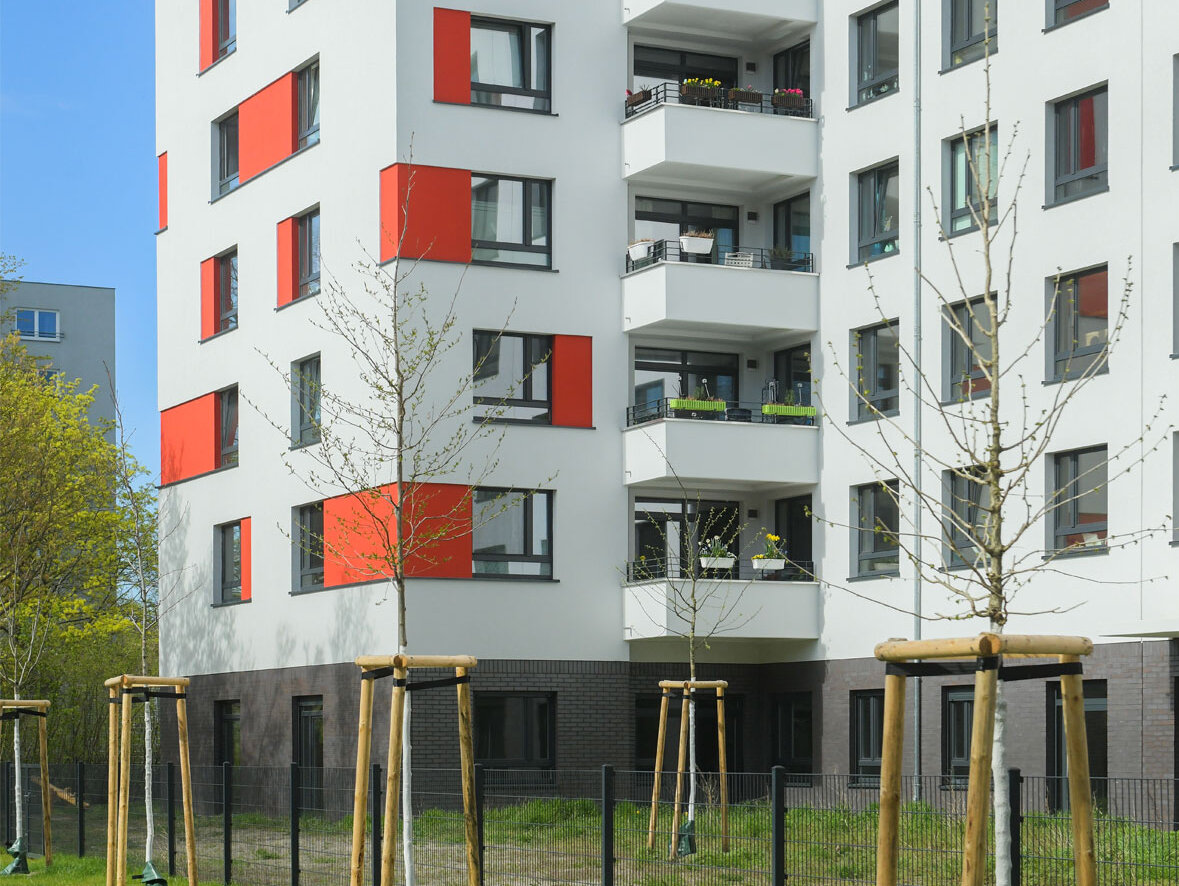 Außenansicht degewo-Neubau An der Wuhlheide 112 bis 116: Hofseite mit Grünflächen, Maschendrahtzaun und neu gepflanzten Bäumen im Vordergrund.
