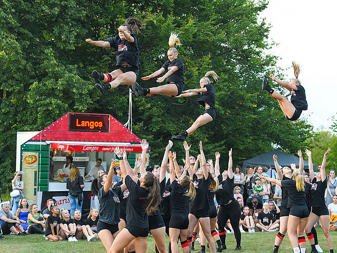 Eine große Gruppe von Cheerleadern zeigt auf einem Fest im Park eine eindrucksvolle Show. Mehrere Mitglieder werden gerade in die Luft geworfen.