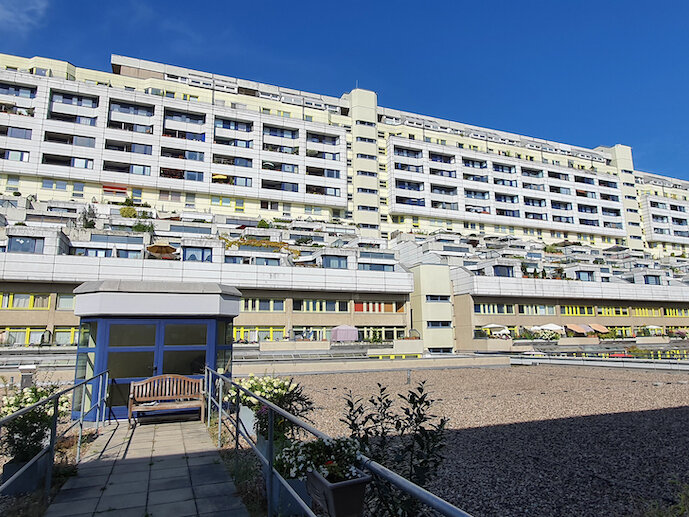 Großes, terrassenförmig angelegtes Wohngebäude mit vielen Balkonen, davor eine Dachfläche und ein kleiner Pavillon unter blauem Himmel.
