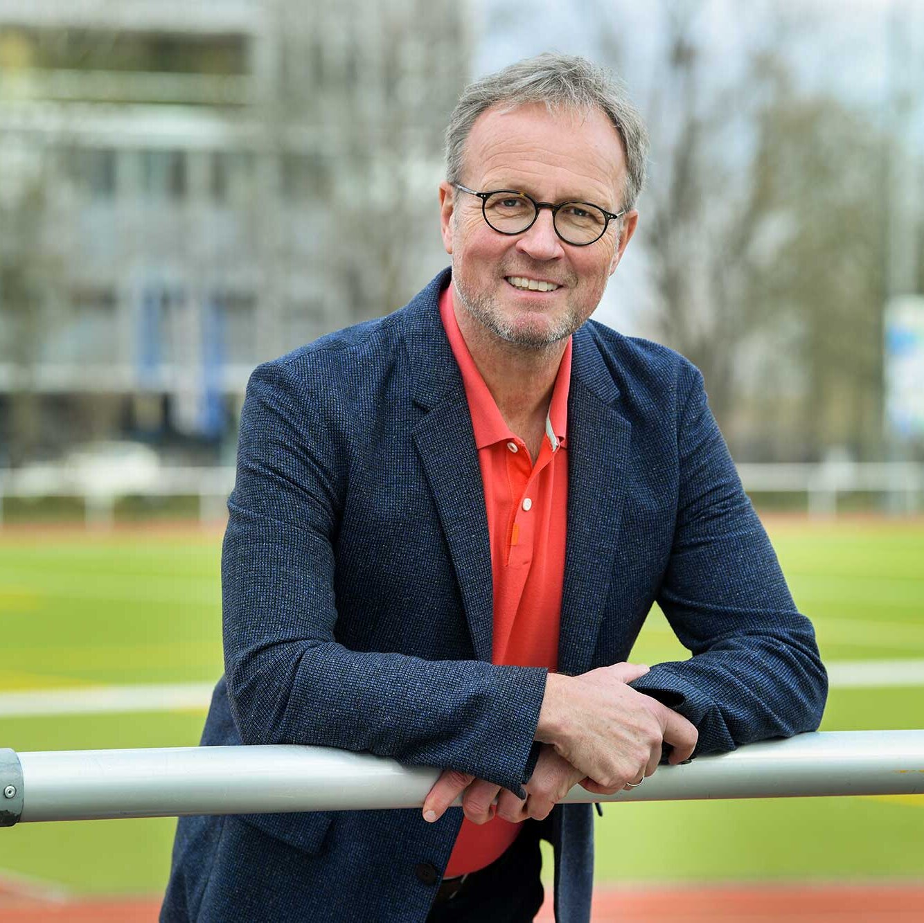 Ein Mann hat seine Arme auf ein Geländer gelegt und lächelt in die Kamera. Im Hintergrund ist verschwommen der Rasen eines Fußballplatzes zu sehen.