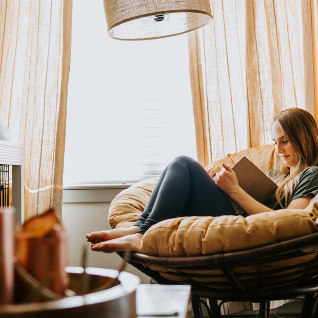 Eine Frau sitzt zum Lesen in einem Sessel am Fenster. Das Zimmer ist mit warmen Farben dekoriert.