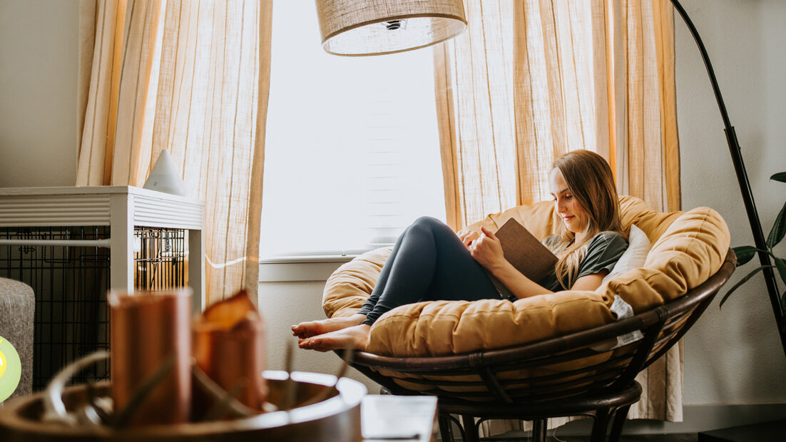 Eine Frau sitzt zum Lesen in einem Sessel am Fenster. Das Zimmer ist mit warmen Farben dekoriert.