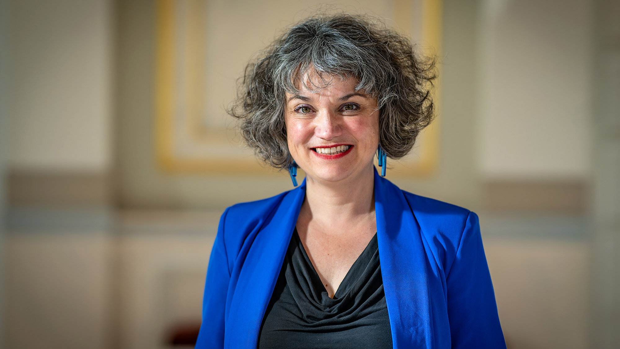 Porträt einer Frau im blauen Sakko vor einem elegant gestalteten Innenraum. Sie lächelt freundlich in die Kamera.