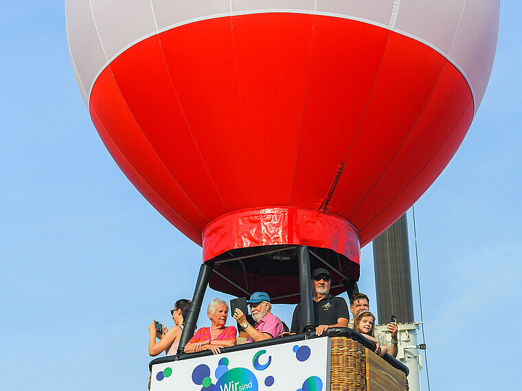 Mehrere Personen befinden sich in einem Heißluftballon. Dieser wird von einem Baukran in den Himmel hinauf gezogen. 