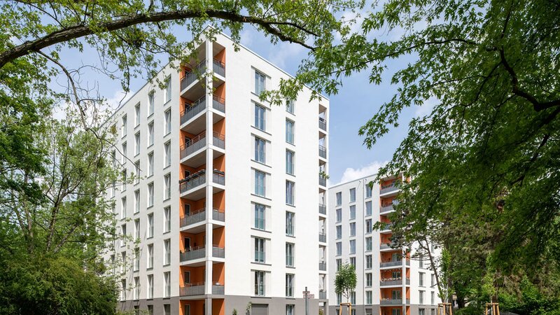 Außenansicht degewo-Neubau Ursulastraße 2 mit Wegen und Rasenflächen sowie links und rechts beblätterten Baumästen und -zweigen im Vordergrund.