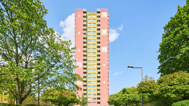 Frontalansicht eines modernisierten degewo-Wohnhochhauses an der Waldsassener Straße in Berlin. Das hohe Gebäude mit rosafarbener Fassade und gelbem Mittelstreifen erhebt sich zwischen grünen Bäumen unter blauem Himmel. Im Vordergrund befinden sich Parkplätze mit abgestellten Autos, die das Wohnumfeld im Quartier zeigen.