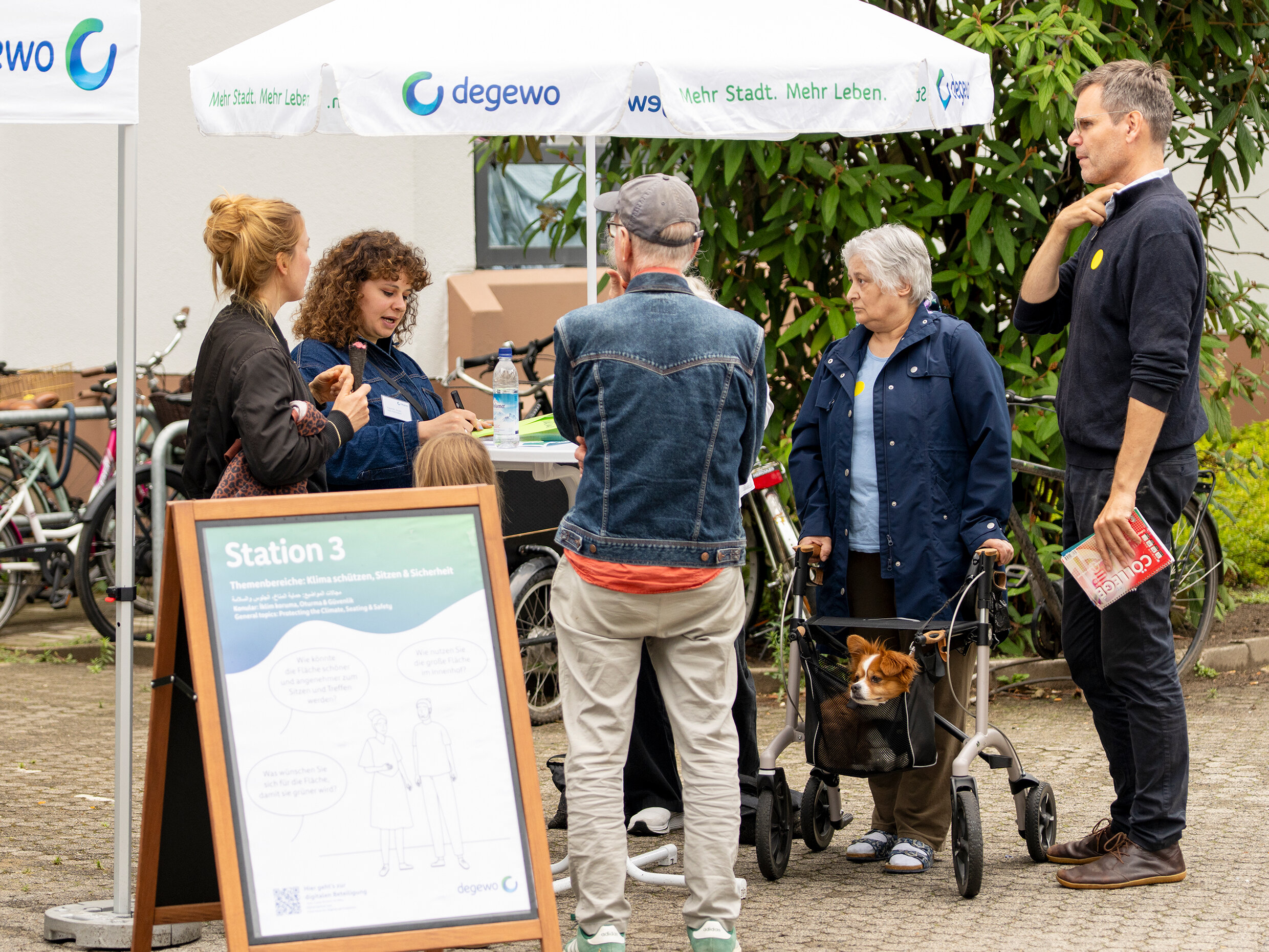Bewohnende im Gespräch an „Station 3“ unter einem degewo-Sonnenschirm. Eine kleine Gruppe diskutiert Ideen zur Gestaltung, ein Hund sitzt im Rollator-Korb einer Teilnehmerin.