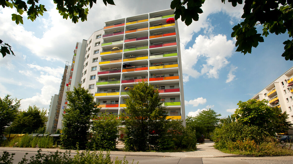 Mehrgeschossiges Wohnhochhaus mit farbigen Balkonbrüstungen in Gelb, Grün und Orange, umgeben von Bäumen und Gehwegen unter blauem Himmel.