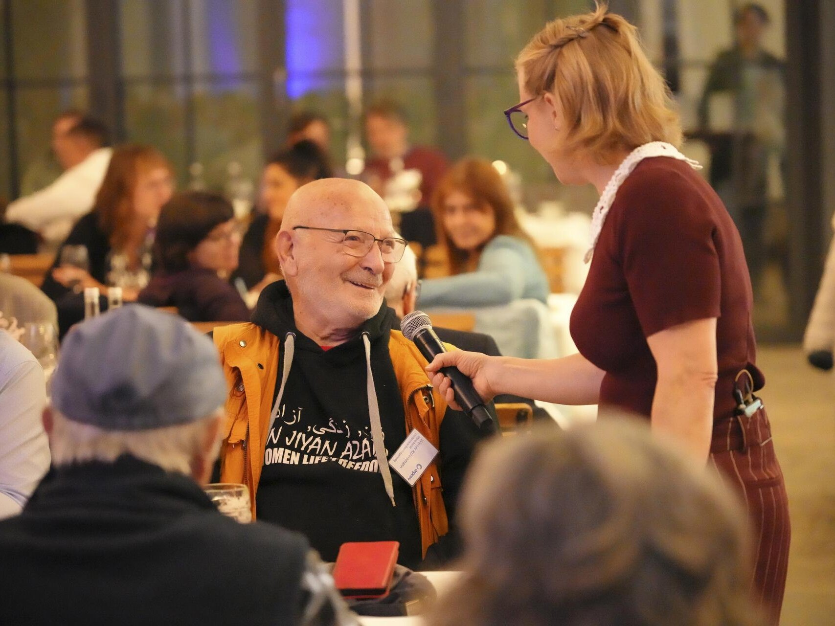 Ein Gast wird von einem Mitglied des Improvisationstheaters interviewt. Er sitzt am Tisch und lacht die Moderatorin an.