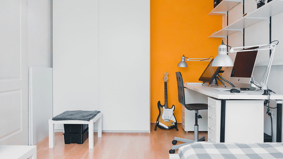 Zimmer mit wenigen Möbeln eingerichtet, an der Wand steht ein Schreibtisch mit Computer.