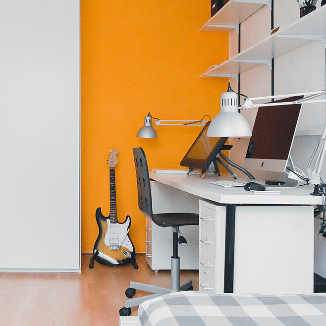 Zimmer mit wenigen Möbeln eingerichtet, an der Wand steht ein Schreibtisch mit Computer.