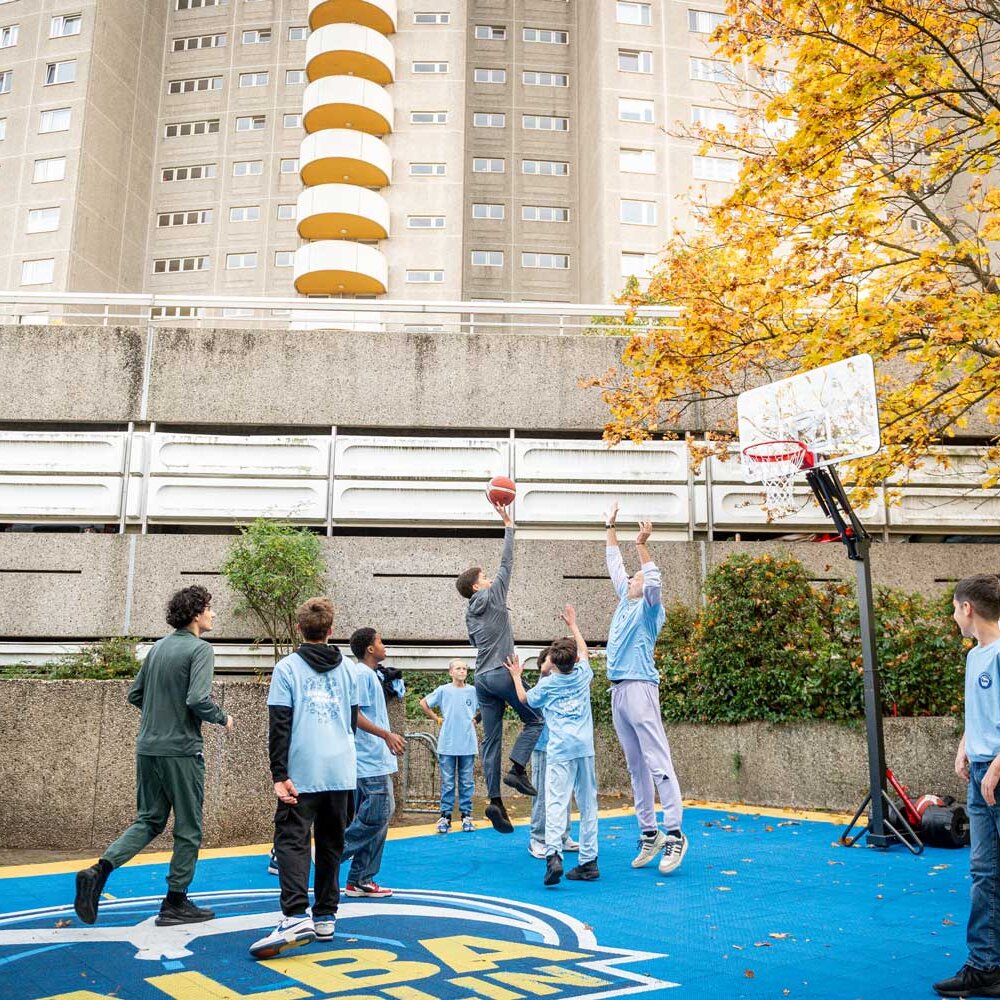 Mehrere Jugendliche spielen Basketball auf einem blau-gelben Outdoor-Court vor einem Wohnhochhaus, während herbstliche Bäume den Platz umrahmen.