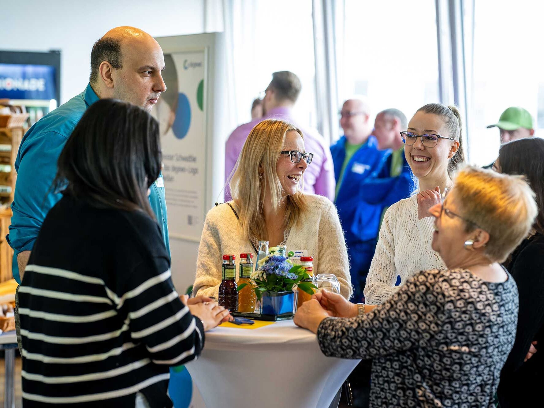 Mehrere Personen stehen an einem Stehtisch bei einer Veranstaltung und unterhalten sich gut gelaunt während Getränke und Blumen auf dem Tisch stehen.