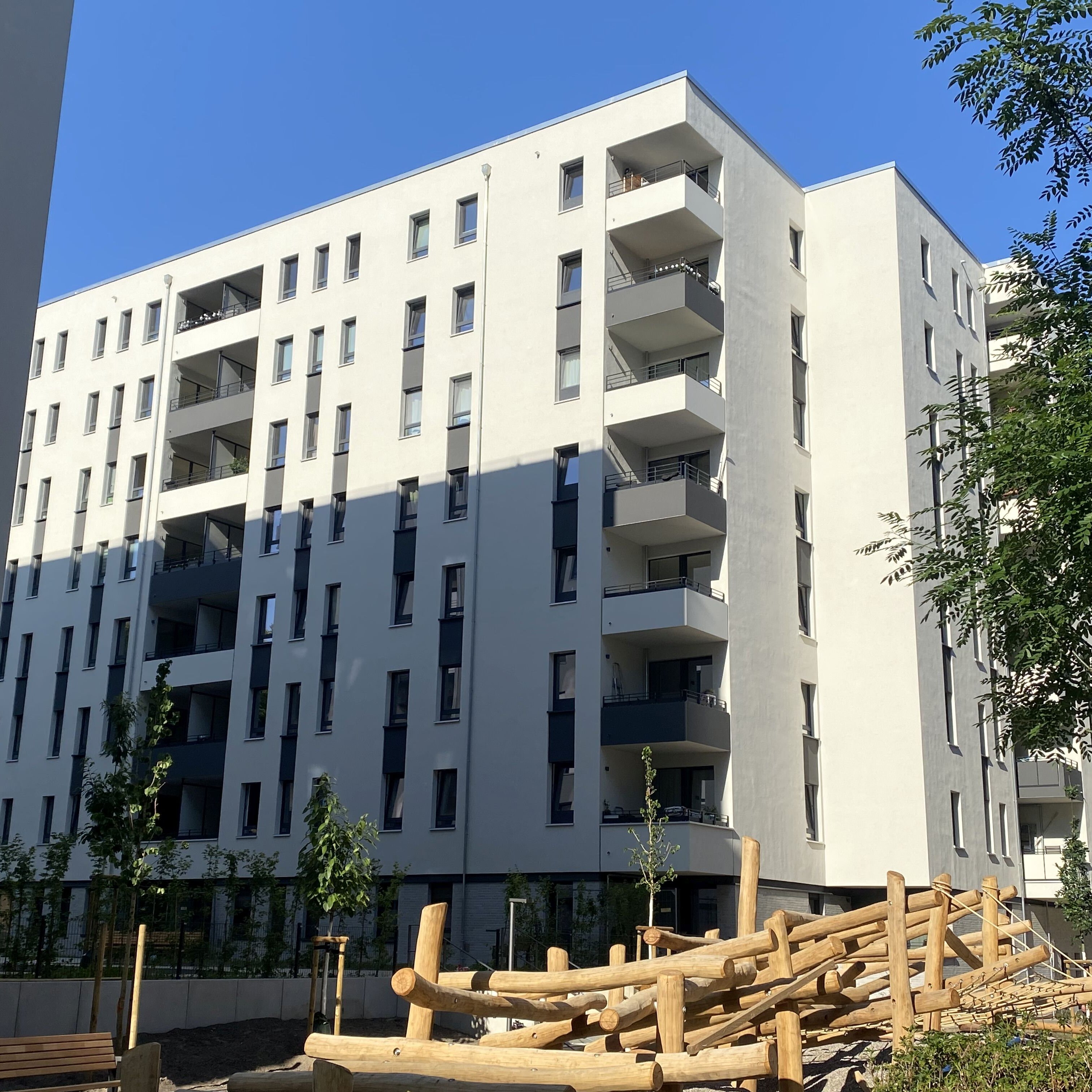 Außenansicht Wohnhaus Hallesches Ufer mit Sitzbank, Klettergerüst, Sträuchern und neu gepflanztem Baum im Vordergrund.