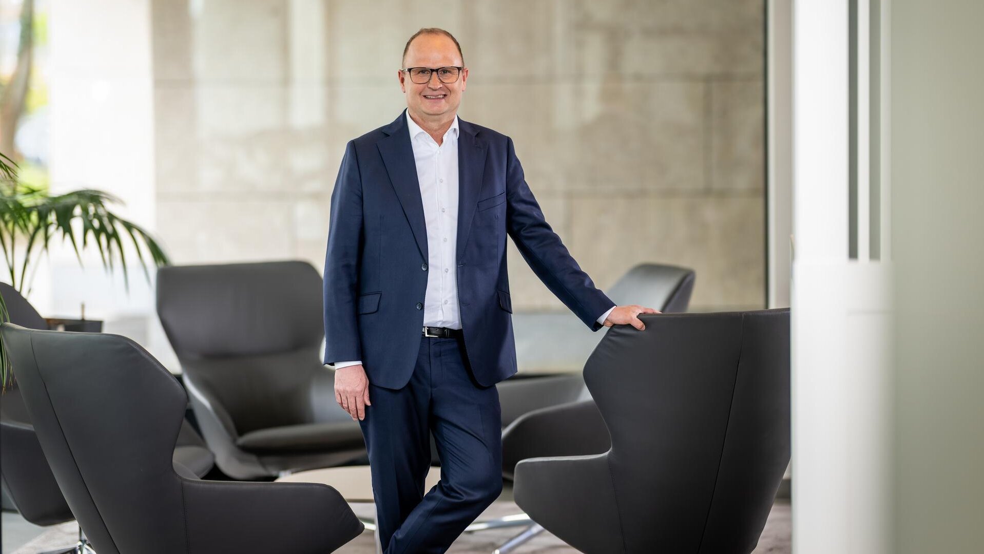 Das Vorstandsmitglied Pascal Atzert steht vor einer Glasscheibe und hat seine Hand auf der Lehne eines Sessels. Er trägt einen Anzug. 