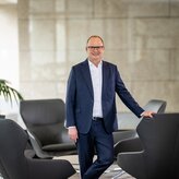 Das Vorstandsmitglied Pascal Atzert steht vor einer Glasscheibe und hat seine Hand auf der Lehne eines Sessels. Er trägt einen Anzug. 