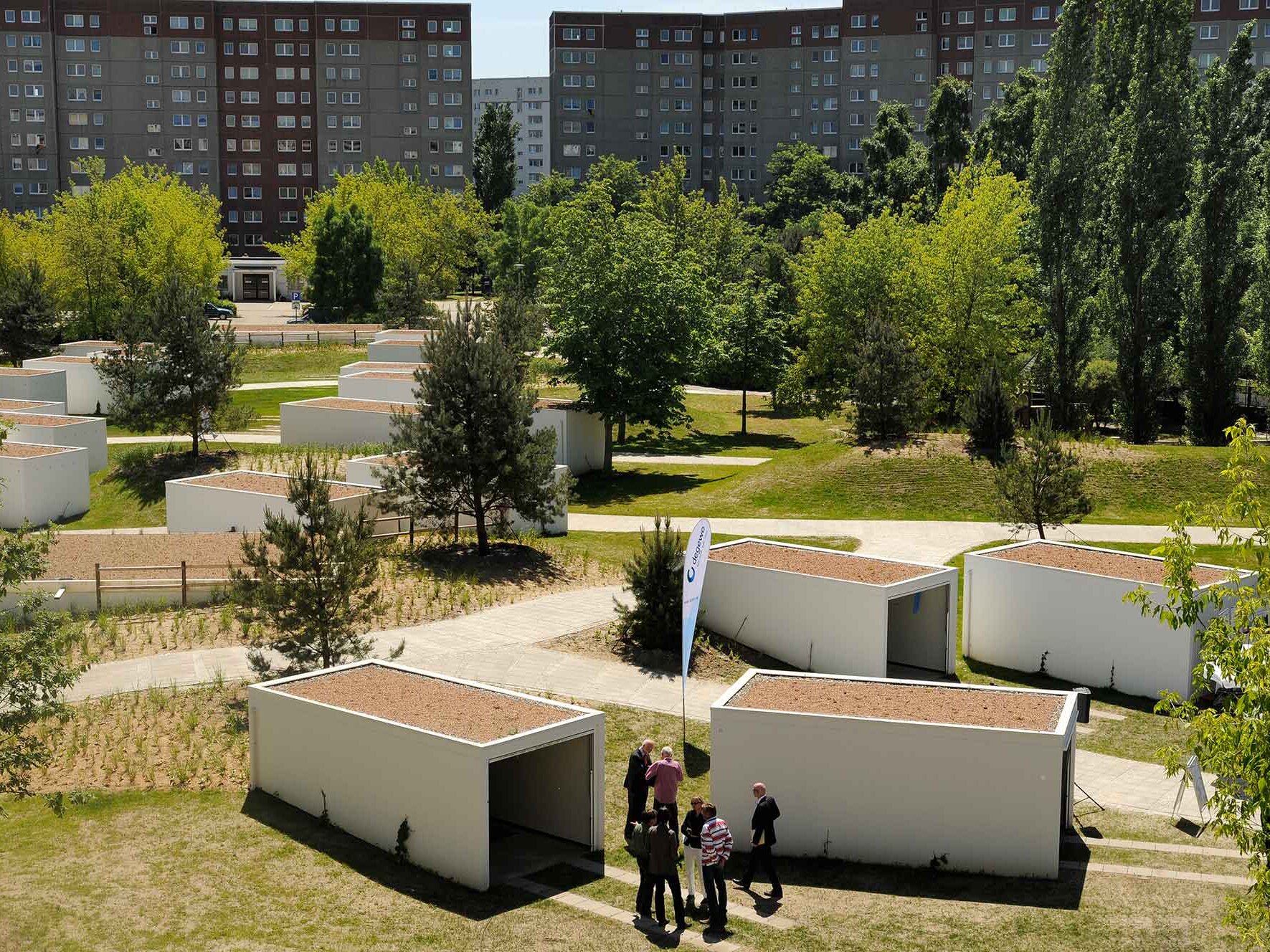 Grünanlage mit mehreren kleinen, weißen, kubusförmigen Gebäuden, zwischen denen Wege verlaufen; im Hintergrund stehen Bäume und große Wohnhäuser.
