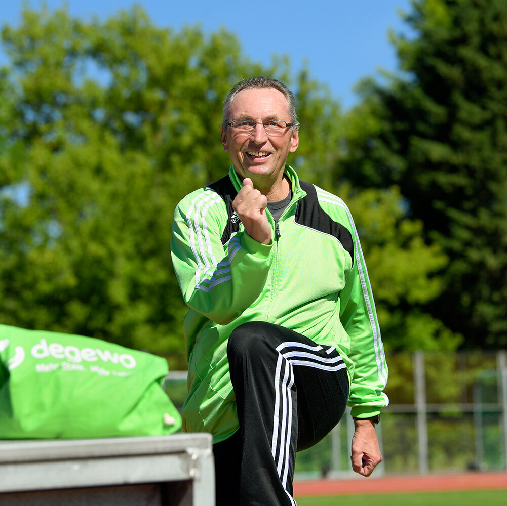 Ein Mann in Sportbekleidung macht auf einem Sportplatz eine Dehnübung. Im Hintergrund grüne Bäume und blauer Himmel.