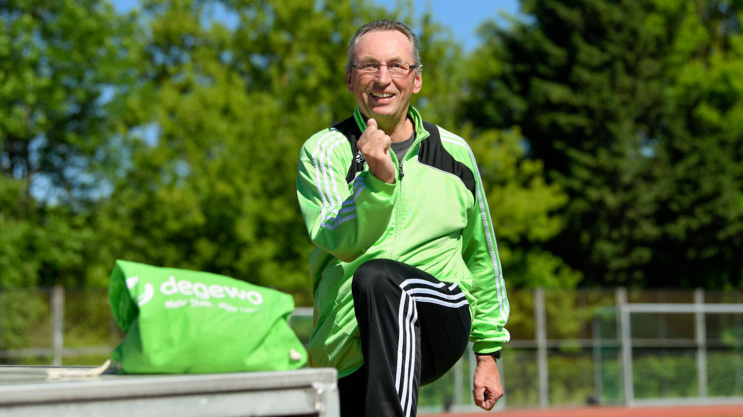 Ein Mann in Sportbekleidung macht auf einem Sportplatz eine Dehnübung. Im Hintergrund grüne Bäume und blauer Himmel.