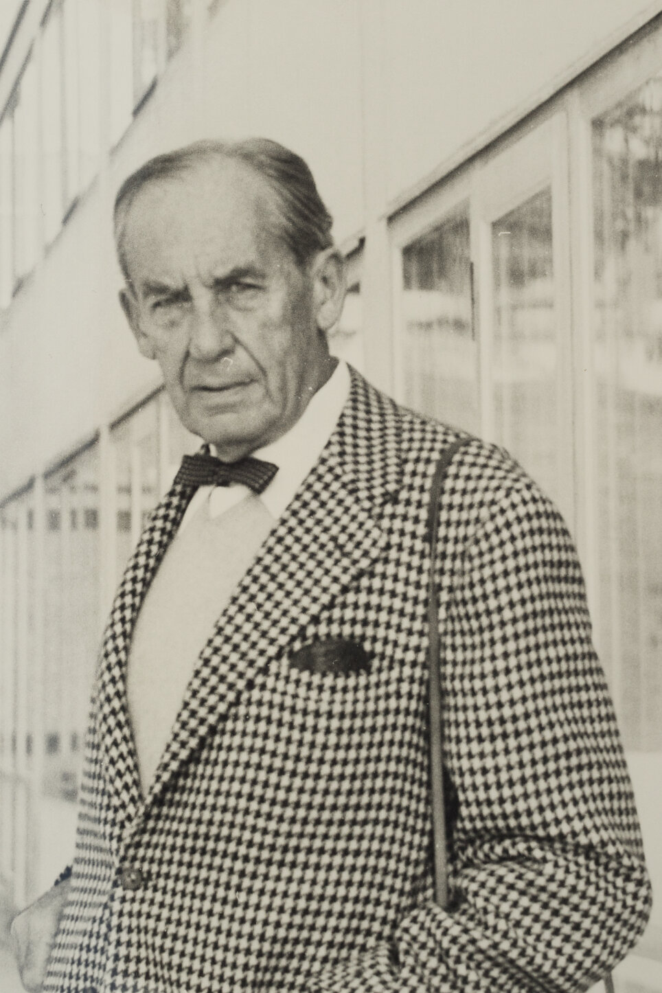 Eine Mann blickt in die Kamera. Er trägt ein kariertes Jacket und eine Fliege. Das Bild ist schwarz-weiß.